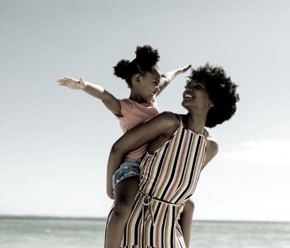 A woman holds a young girl on her hip by the beach; the girl extends her arms out as if to fly, while both appear to be smiling and enjoying the moment after a peaceful resolution through Fair Divorce Solutions